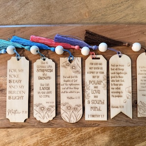 May include: Wooden bookmarks with inspirational quotes and Bible verses. Each bookmark has a colorful tassel and a wooden bead. The quotes are engraved in a clear, readable font. The bookmarks are arranged on a wooden surface.