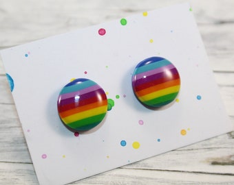 Könnte beinhalten: Zwei runde Ohrstecker mit Regenbogenstreifen auf einer weißen Karte mit bunten Farbspritzern. Die Ohrringe haben eine glänzende Oberfläche und weisen Bänder in Blau, Lila, Rot, Gelb und Grün auf. Die Karte ist leicht angewinkelt.