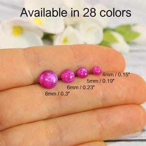 May include: A hand holds five pink, glittery, round studs in different sizes: 4mm (0.15"), 5mm (0.19"), 6mm (0.23"), and 8mm (0.3"). The text "Available in 28 colours" is at the top. The studs are set against a blurred background.