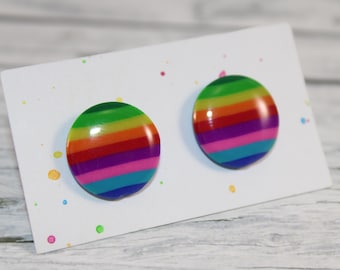 Könnte beinhalten: Ein Paar runde Ohrstecker mit einem Regenbogen-Streifen-Design. Die Ohrringe werden auf einer weißen Karte mit bunten Farbspritzern präsentiert. Die Ohrringe haben einen Durchmesser von etwa 2,5 cm.