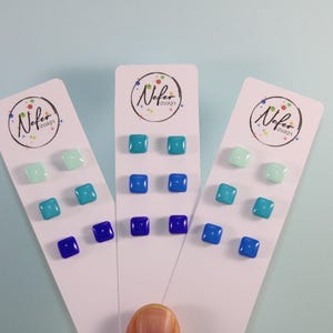 Op de afbeelding: Drie witte displaykaarten, elk voorzien van het Nefer design logo, bevatten paren vierkante oorknopjes in verschillende blauwtinten. De oorbellen variëren van licht muntgroen tot turquoise, hemelsblauw en diep koningsblauw in een kleurverloop.