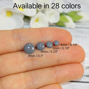 Peut inclure: Quatre perles grises de différentes tailles, de 4 mm à 8 mm (0,15" à 0,3"), sont présentées. Le texte "Disponible en 28 couleurs" est en haut. Les perles sont présentées sur un fond flou de fleurs blanches.