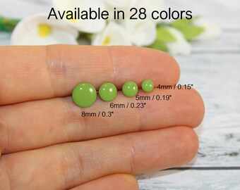 Op de afbeelding: Vier groene oorstekers in verschillende maten, van 4 mm tot 8 mm (0,3 inch), worden getoond. De tekst "Verkrijgbaar in 28 kleuren" staat bovenaan. De oorbellen worden in een hand gehouden.