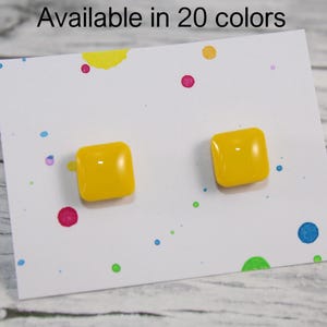Puede incluir: Un par de pendientes cuadrados amarillos brillantes sobre una tarjeta blanca con salpicaduras de pintura de colores. El texto "Available in 20 colors" está en la parte superior. Los pendientes son brillantes y tienen esquinas redondeadas.