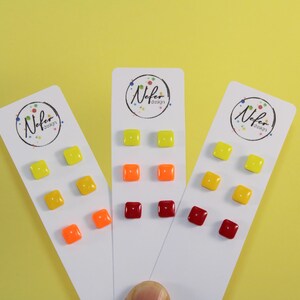 Op de afbeelding: Drie witte rechthoekige displaykaarten, elk met het Nefer Design-logo, bevatten paren kleine vierkante oorknopjes in levendige tinten geel, oranje en donkerrood. De oorknopjes hebben een glanzende afwerking en zijn in rijen gerangschikt.