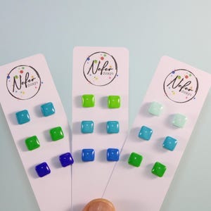 Op de afbeelding: Drie witte presentatiekaarten, elk met het ronde Nefer design logo, bevatten paren kleine, vierkante, glanzende oorknopjes in diverse tinten blauw en groen, waaronder lichtblauw, heldergroen, donkerblauw en licht mintgroen.
