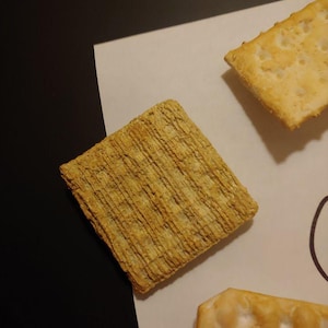 May include: Close-up of three golden-brown crackers with a textured surface. One cracker is square, while the other two are rectangular. The crackers are on a white surface with a black background.