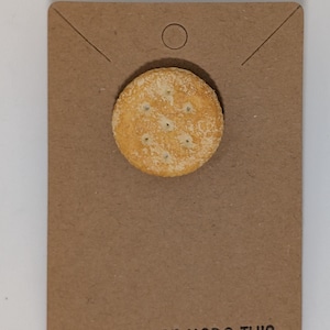 May include: A round, light brown cracker-shaped pin is affixed to a brown card. The pin has small holes and a textured surface. The card has a circular hole at the top and the text "SUE MILLER MADE THIS" printed at the bottom.