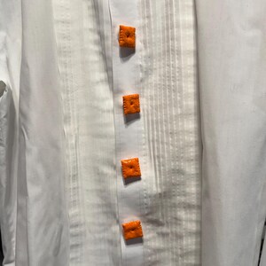 May include: White dress shirt with a pleated front and six square, orange, textured buttons. The buttons are evenly spaced down the center of the shirt.