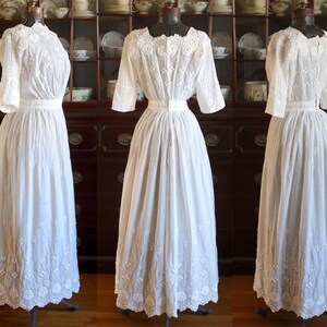 May include: Three white vintage dresses on display. Each dress features a high neckline, short sleeves, and a long skirt. The dresses are made of a delicate fabric with intricate embroidery details. The dresses are displayed on mannequins.