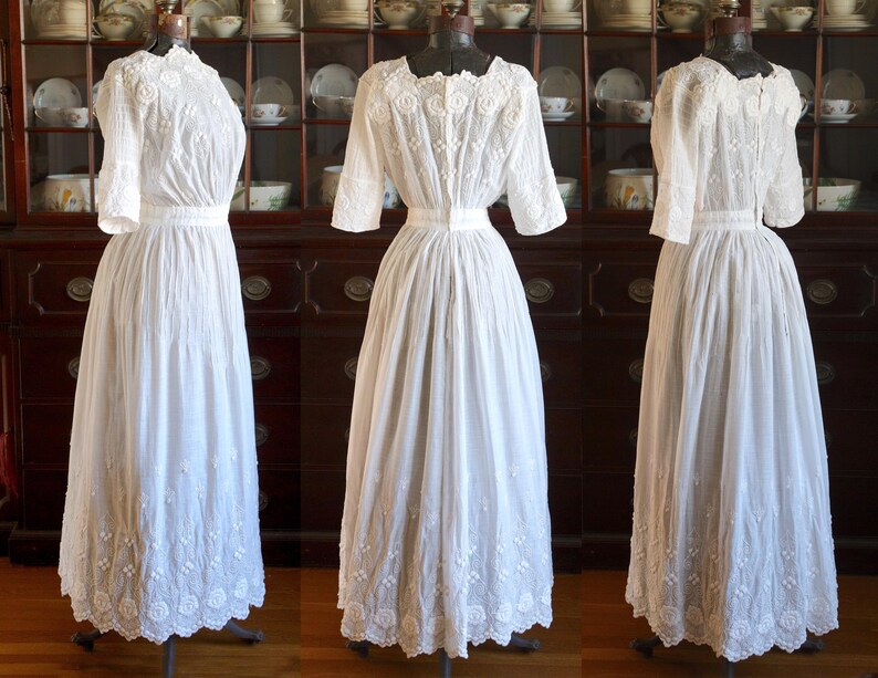 May include: Three white vintage dresses on display. Each dress features a high neckline, short sleeves, and a long skirt. The dresses are made of a delicate fabric with intricate embroidery details. The dresses are displayed on mannequins.