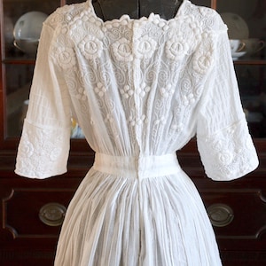 May include: A white vintage dress with intricate floral embroidery on the bodice and sleeves. The dress features a square neckline, short sleeves, and a pleated skirt. The back of the dress is buttoned up.