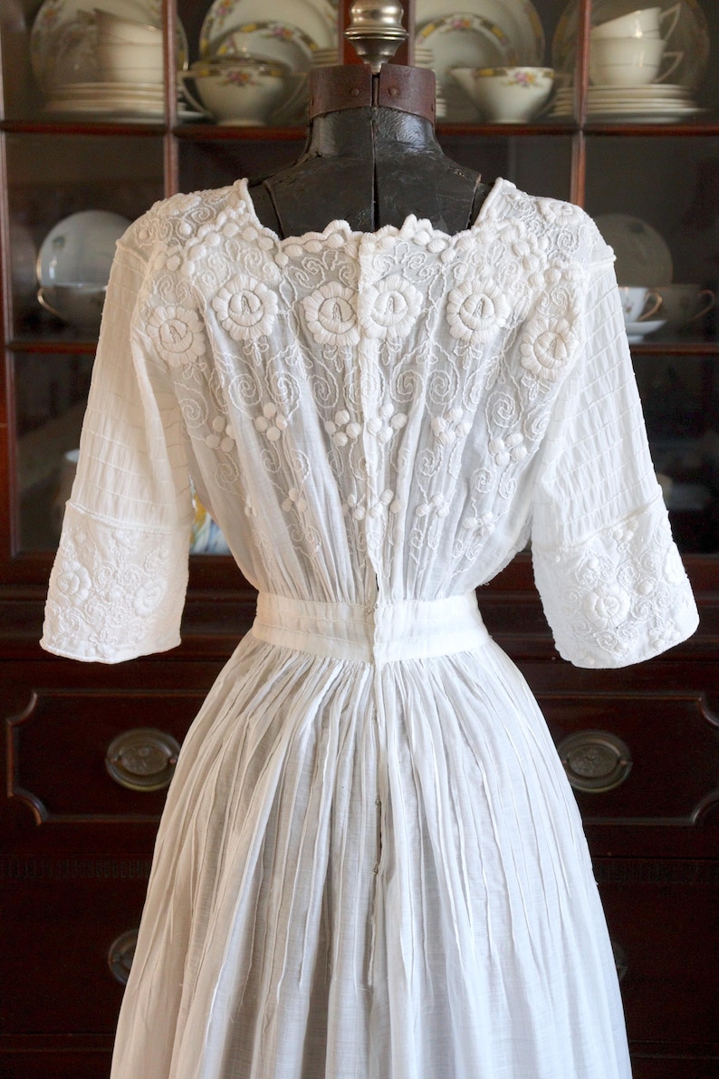 May include: A white vintage dress with intricate floral embroidery on the bodice and sleeves. The dress features a square neckline, short sleeves, and a pleated skirt. The back of the dress is buttoned up.
