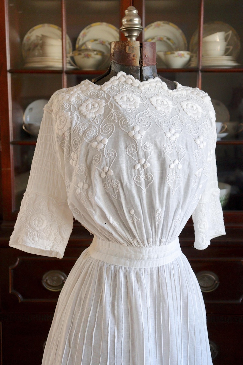 May include: A white vintage dress with intricate floral embroidery on the bodice and sleeves. The dress features a fitted waist and a pleated skirt, displayed on a vintage dress form. The background includes a wooden cabinet with antique dishware.