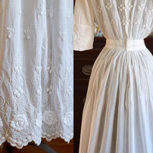 May include: A white, vintage dress with intricate floral and scroll embroidery. The dress features short sleeves, a high neckline, and a fitted waist. The hemline is detailed with a scalloped edge. The fabric appears to be lightweight and breathable.