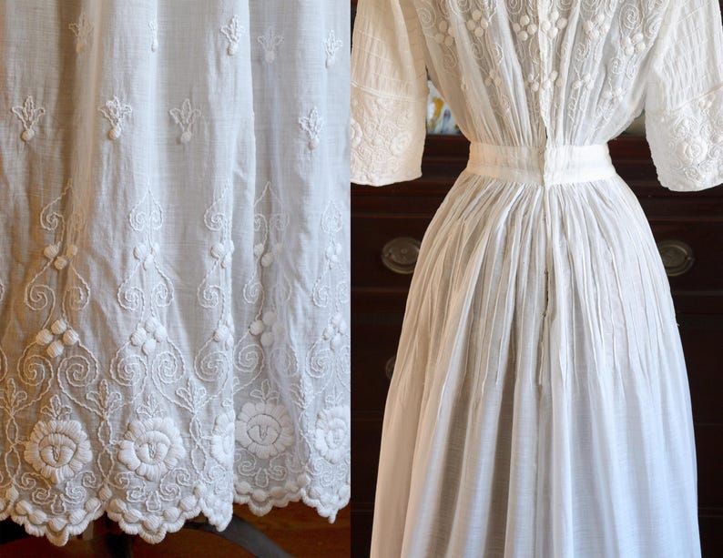 May include: A white, vintage dress with intricate floral and scroll embroidery. The dress features short sleeves, a high neckline, and a fitted waist. The hemline is detailed with a scalloped edge. The fabric appears to be lightweight and breathable.