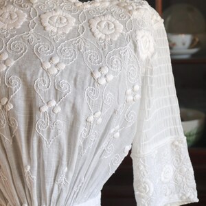 May include: A white vintage dress with intricate floral embroidery. The dress features a high neckline, long sleeves, and a fitted waist. The embroidery includes detailed floral patterns and delicate scrollwork, creating a romantic and elegant look.
