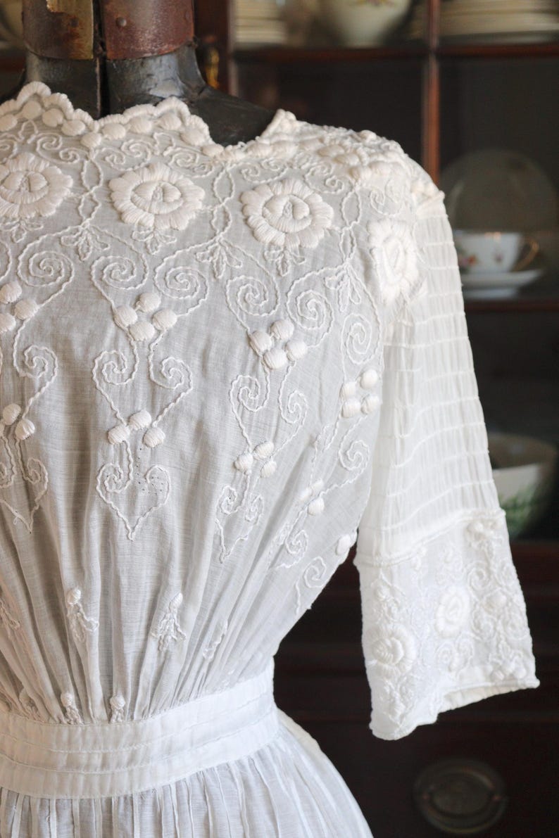 May include: A white vintage dress with intricate floral embroidery. The dress features a high neckline, long sleeves, and a fitted waist. The embroidery includes detailed floral patterns and delicate scrollwork, creating a romantic and elegant look.