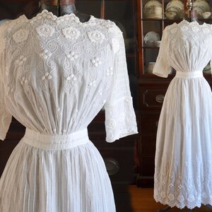 May include: A white, vintage-style dress with intricate floral embroidery on the bodice and sleeves. The dress features a high neckline, a fitted waist, and a pleated skirt. The sleeves are three-quarter length. The dress is displayed on a mannequin.