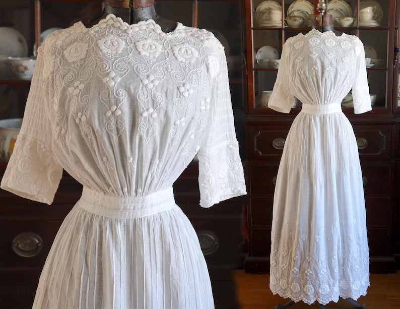 May include: A white, vintage-style dress with intricate floral embroidery on the bodice and sleeves. The dress features a high neckline, a fitted waist, and a pleated skirt. The sleeves are three-quarter length. The dress is displayed on a mannequin.