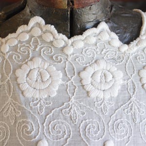 May include: Close-up of a white embroidered garment. The fabric features intricate floral and scrollwork patterns, with a scalloped neckline. The embroidery is detailed and raised, creating a textured surface. The garment is displayed on a mannequin.