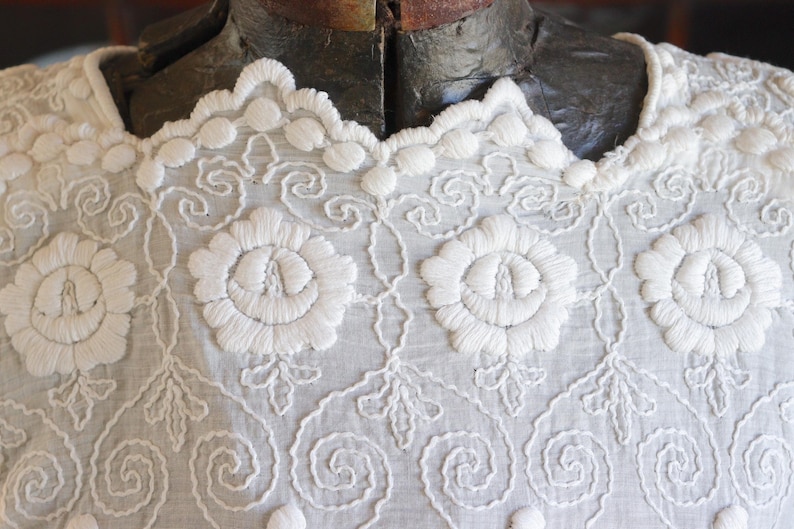 May include: Close-up of a white embroidered garment. The fabric features intricate floral and scrollwork patterns, with a scalloped neckline. The embroidery is detailed and raised, creating a textured surface. The garment is displayed on a mannequin.