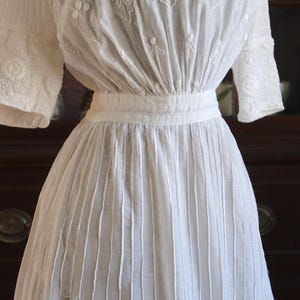 May include: A white vintage dress with intricate floral embroidery on the bodice and sleeves. The dress features a fitted waist and a pleated skirt, creating a classic silhouette. The fabric appears to be lightweight, suitable for warmer weather.