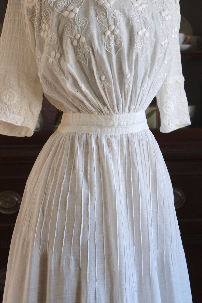May include: A white vintage dress with intricate floral embroidery on the bodice and sleeves. The dress features a fitted waist and a pleated skirt, creating a classic silhouette. The fabric appears to be lightweight, suitable for warmer weather.