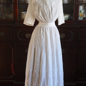 May include: A vintage white dress with intricate floral embroidery. The dress features a high neckline, short sleeves, and a fitted waist. The skirt is long and flowing, with detailed embroidery along the hemline. The dress is displayed on a mannequin.