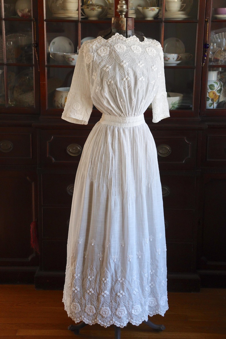 May include: A vintage white dress with intricate floral embroidery. The dress features a high neckline, short sleeves, and a fitted waist. The skirt is long and flowing, with detailed embroidery along the hemline. The dress is displayed on a mannequin.