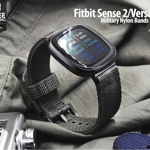 Pode incluir: Um smartwatch Fitbit Sense 2/Versa 4 preto com uma pulseira de nylon verde escura. O mostrador do relógio mostra a hora, a data, os passos, a distância e a frequência cardíaca.