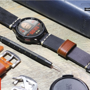 May include: A black leather watch band with brown leather accents and white stitching. The watch face is black with a white digital display showing the time and date. The text "THE PRIMRIA WATCH BANDS & STRAPS" is visible on a blue leather case.