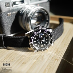 May include: A black and silver Rolex Submariner watch with a black nylon strap. The watch has a black dial with white markings and a date window at the 3 o'clock position. The watch is on a wooden surface.