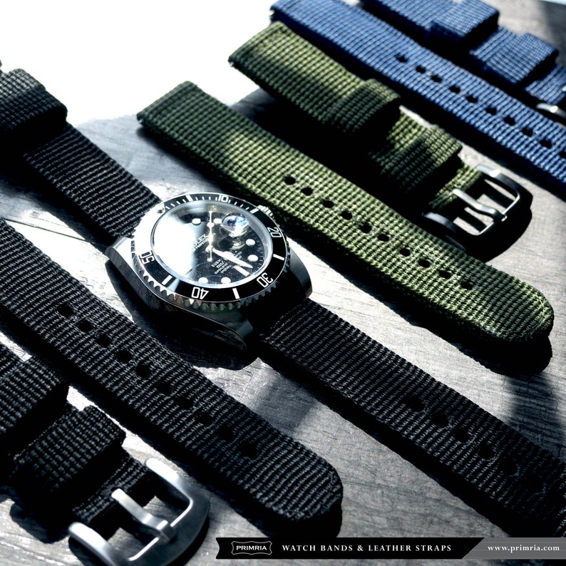 May include: A close-up of a black and silver watch with a black nylon watch strap. The watch is surrounded by other nylon watch straps in various colors, including green and blue.
