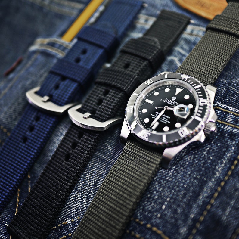 May include: A black Rolex Submariner watch with a black face and silver hands and markers. The watch is on a green nylon strap and is laying on top of a blue denim fabric.