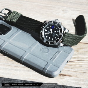 May include: A black and silver watch with a green nylon strap. The watch has a black face with white markings and a date window. The strap is attached to the watch with silver lugs. The watch is lying on a gray phone case with the words "RUGGED SHIELD" printed on it.