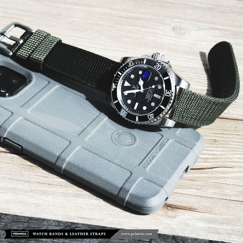 May include: A black and silver watch with a green nylon strap. The watch has a black face with white markings and a date window. The strap is attached to the watch with silver lugs. The watch is lying on a gray phone case with the words "RUGGED SHIELD" printed on it.