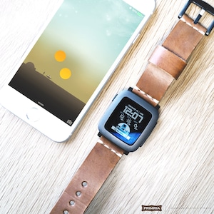 K&ouml;nnte beinhalten: Ein braunes Lederarmband mit einem schwarzen Smartwatch-Zifferblatt, das die Zeit 12:07 und das Datum Donnerstag, 27. August anzeigt. Das Zifferblatt zeigt auch ein Wettersymbol und eine Temperatur von 25 Grad an. Die Smartwatch liegt auf einer Holzoberfl&auml;che neben einem wei&szlig;en iPhone mit einem Star Wars-Hintergrundbild.