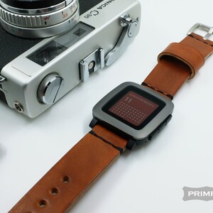 K&ouml;nnte beinhalten: Ein braunes Lederarmband mit einer silbernen Pebble Smartwatch. Das Zifferblatt der Uhr zeigt das Datum und den Wochentag an, "WEDNESDAY 11".