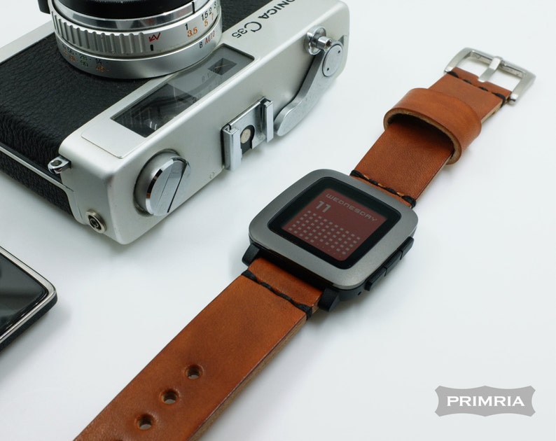 K&ouml;nnte beinhalten: Ein braunes Lederarmband mit einer silbernen Pebble Smartwatch. Das Zifferblatt der Uhr zeigt das Datum und den Wochentag an, "WEDNESDAY 11".