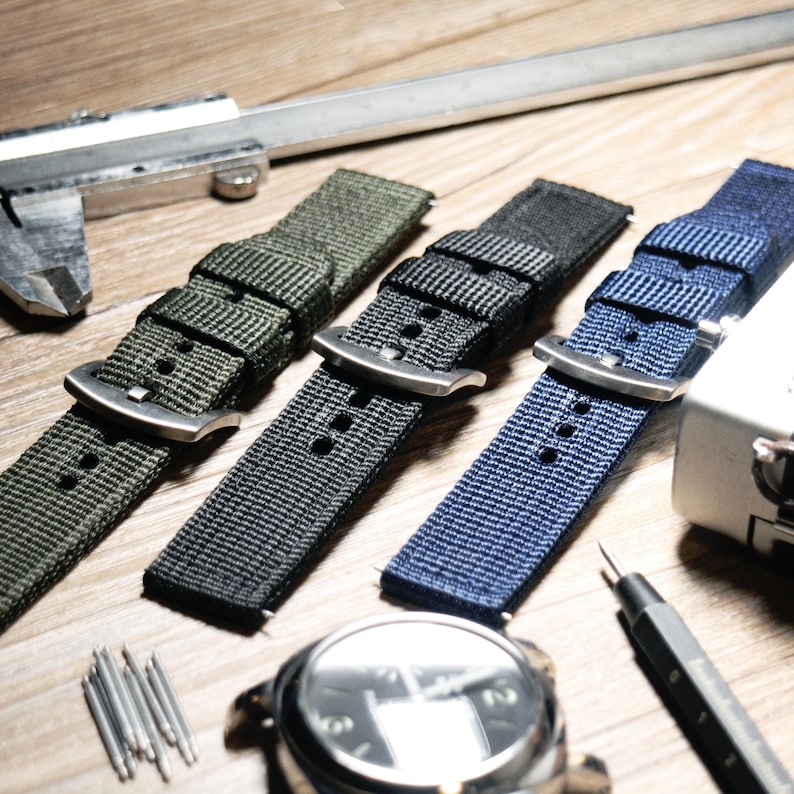 May include: Three different colored nylon watch straps with silver buckles. The straps are green, black, and blue. The straps are laid out on a wooden surface.