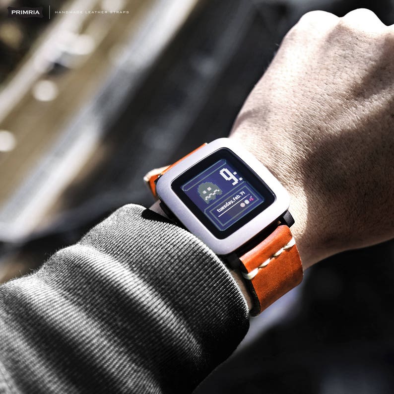 K&ouml;nnte beinhalten: Eine silberne Pebble Smartwatch mit einem braunen Lederarmband. Das Zifferblatt zeigt die Uhrzeit, das Datum und einen Pac-Man-Charakter.