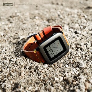 K&ouml;nnte beinhalten: Eine graue Pebble Smartwatch mit einem schwarzen Lederarmband. Das Zifferblatt der Uhr zeigt die Zeit, das Datum und die Temperatur an. Die Uhr liegt auf einem Bett aus kleinen grauen Steinen.
