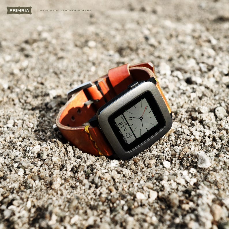 K&ouml;nnte beinhalten: Eine graue Pebble Smartwatch mit einem schwarzen Lederarmband. Das Zifferblatt der Uhr zeigt die Zeit, das Datum und die Temperatur an. Die Uhr liegt auf einem Bett aus kleinen grauen Steinen.
