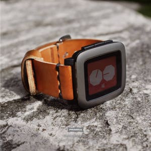 K&ouml;nnte beinhalten: Ein braunes Lederarmband mit einer silbernen Pebble Time Smartwatch. Das Zifferblatt der Uhr zeigt die Zeit im digitalen Format an.