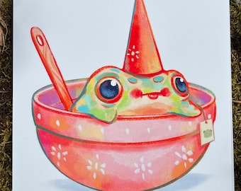 May include: A colourful frog wearing a red cone-shaped hat sits in a pink bowl with a wooden spoon and a tea bag. The bowl has a white floral pattern.
