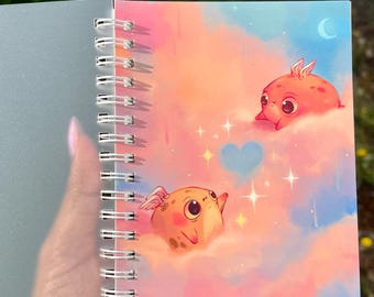 Puede incluir: Un cuaderno de espiral con una ilustración en la portada de dos pequeñas criaturas aladas de color naranja que flotan entre nubes rosas y azules. El diseño incluye un corazón azul claro, estrellas brillantes y una luna creciente.