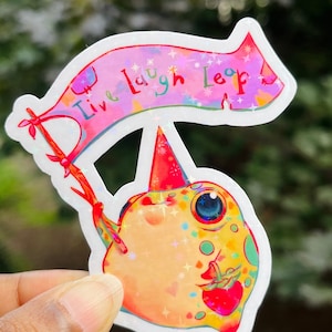 May include: A sticker featuring a cartoon fish wearing a party hat and holding a banner that says "Live Laugh Leap". The fish is pink, yellow, and green with a red strawberry in its mouth. The banner is pink with rainbow glitter.