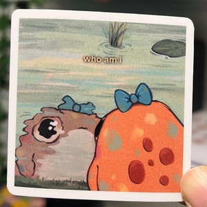 May include: A sticker with a cartoon image of two characters, one brown and one orange, with blue bows on their heads. The text "who am i" is written in the background.