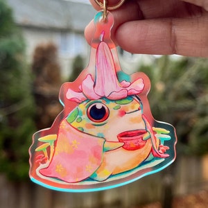 May include: A colorful acrylic keychain featuring a whimsical frog character. The frog is adorned with a flower hat and a pink shawl, holding a teacup. The keychain has a gold-colored ring.